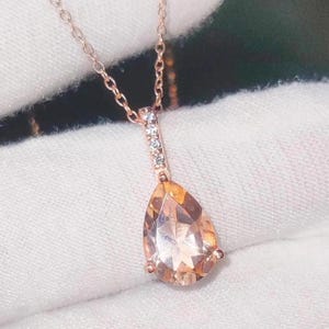 Puede incluir: Un collar de oro rosa con un colgante de piedra preciosa en forma de pera, color melocotón. El colgante está adornado con pequeñas piedras preciosas transparentes. La cadena y el engaste tienen un diseño delicado.