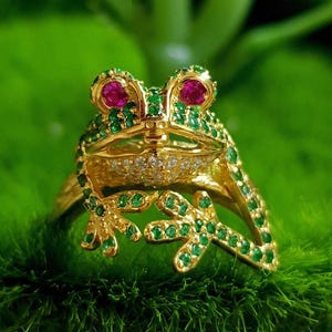 Puede incluir: Un anillo dorado con forma de rana, adornado con piedras preciosas verdes y rojas. Los ojos de la rana son rojos y su cuerpo está cubierto de pequeñas piedras verdes. La banda del anillo presenta las patas de la rana, también adornadas con piedras verdes.