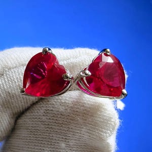 Può includere: Un paio di orecchini a forma di cuore con gemme rosse vibranti. Gli orecchini sono incastonati in metallo color argento e sono esposti su uno sfondo di tessuto bianco con uno sfondo blu.