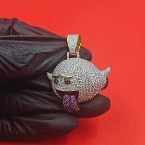 May include: A gold-toned pendant in the shape of a cartoon ghost, covered in small, sparkling stones. The ghost has black eyes, a gold mustache, and a purple tongue. The pendant is held against a red background.