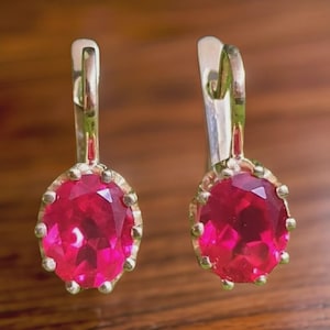 Könnte beinhalten: Ein Paar goldfarbene Ohrringe mit großen, oval geschliffenen, leuchtend roten Edelsteinen. Die Edelsteine sind in einer goldfarbenen Lünette mit kleinen, dekorativen Akzenten gefasst. Mit Klappbügelverschluss.