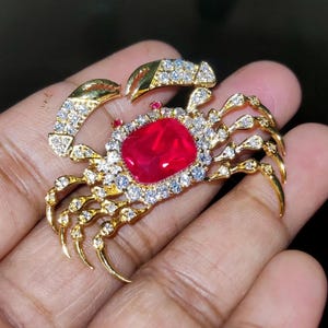 Pode incluir: Broche de caranguejo dourado com uma grande pedra vermelha no centro. As garras e o corpo do caranguejo são adornados com cristais transparentes e brilhantes. Este broche detalhado é uma joia ornamentada.