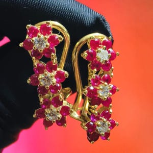 Könnte beinhalten: Ein Paar goldene Ohrringe mit floralem Design. Jeder Ohrring hat drei Blumencluster mit leuchtend roten Blütenblättern und einem zentralen weißen Edelstein. Die Ohrringe haben ein gebogenes, hakenförmiges Design.