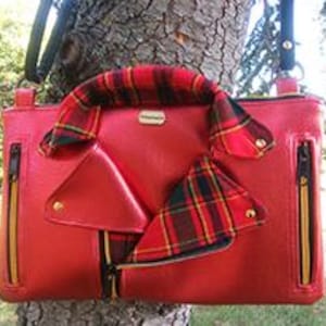 May include: A red handbag with a unique design resembling a biker jacket. The bag features a plaid handle and accents, with gold-colored zippers and hardware. The bag is shown against a tree trunk.