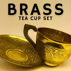 May include: A brass tea cup set with a matching saucer. The cup and saucer are gold-colored and feature intricate floral patterns. The cup has a curved handle and the text "KACHI RASOI" is visible inside the cup. The words "BRASS TEA CUP SET" are at the top.