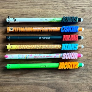 Könnte beinhalten: Eine Sammlung farbenfroher Stifte mit Tiermotiven und Namen. Die Stifte zeigen Sätze wie "HUG ME" und "BELIEVE IN MAGIC". Jeder Stift hat einen anderen Namen in fetten Buchstaben, darunter Jonas, Oskar, Hilma, Klaas, Sonja und Moritz.