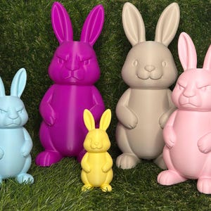 Könnte beinhalten: Eine Sammlung farbenfroher Hasenfiguren in verschiedenen Größen. Die Hasen sind in Blau-, Lila-, Gelb-, Beige- und Rosatönen gehalten. Einige Hasen haben einen strengen Ausdruck, andere wirken freundlicher. Die Figuren stehen auf grünem Gras.