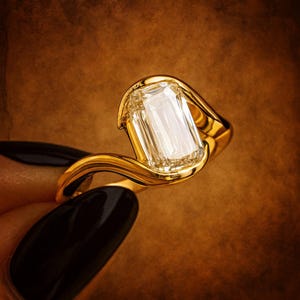 Op de afbeelding: Een gouden ring met een grote, rechthoekig geslepen diamant. De ringband heeft een vloeiend, gebogen ontwerp. De diamant is helder en reflecteert licht, tegen een warme bruine achtergrond. De ring is een sieraad.