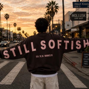 May include: A person stands in a crosswalk wearing a brown sweatshirt with the words "STILL SOFTISH" in pink across the back. The sweatshirt also has the Roman numerals "III/XX/MMXX". The background features a street scene with palm trees and storefronts.
