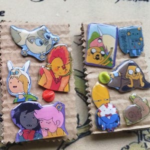 Può includere: Una collezione di spille smaltate colorate raffiguranti personaggi dei cartoni animati, disposte su un supporto di cartone. Le spille presentano vari disegni, tra cui figure con colori vivaci e opere d'arte dettagliate. Le spille sono fissate con chiusure a spillo.