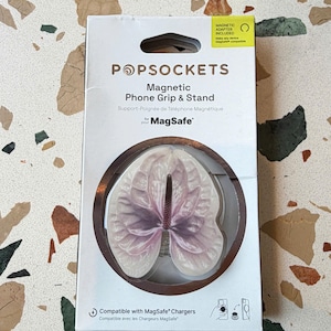 May include: A white retail box for a PopSockets magnetic phone grip and stand. The packaging features a graphic of a pale pink and white anthurium flower. Text on the box includes PopSockets, Magnetic Phone Grip & Stand, and MagSafe.