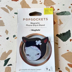 以下が含まれることがあります： 白いパッケージに入ったPopSocketsの磁気式スマホグリップ兼スタンド。黒い耳、白い顔、頭に絆創膏を貼ったキャラクターのグラフィックが特徴です。パッケージには「Magnetic Adapter Included」および「Compatible with MagSafe Chargers」というテキストが記載されています。