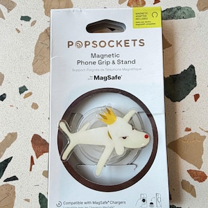 May include: A white PopSockets magnetic phone grip and stand in its original retail packaging. The product features a cartoon dog wearing a yellow crown. The packaging includes text stating Magnetic Phone Grip & Stand for MagSafe and Magnetic Adapter Included.