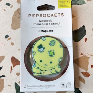 May include: A PopSockets magnetic phone grip and stand in its retail packaging. The product features a pale yellow circular design with a cartoon character writing in a notebook, surrounded by small icons. The packaging includes the text PopSockets, Magnetic Phone Grip & Stand, and MagSafe compatibility information.