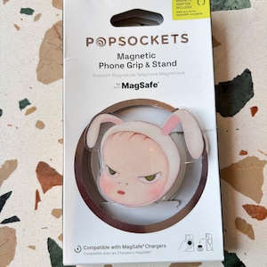 Op de afbeelding: Een magnetische PopSockets telefoongreep en standaard in een witte verpakking. Het product heeft een rond ontwerp met een stripfiguur die een muts met konijnenoren draagt. De verpakking bevat de tekst PopSockets, Magnetic Phone Grip & Stand en MagSafe.