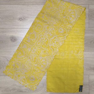 May include: A rectangular scarf featuring a bright yellow and cream-coloured geometric pattern on one half and a solid, textured yellow fabric on the other half. The scarf has a small black rectangular tag attached to the bottom right corner.