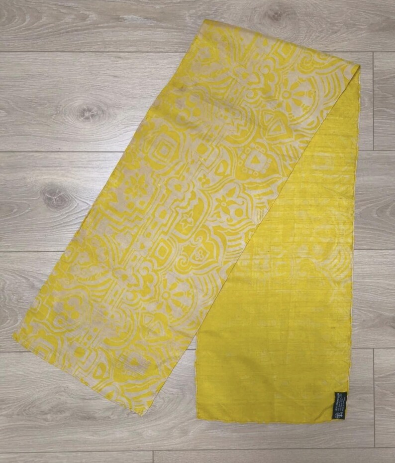 May include: A rectangular scarf featuring a bright yellow and cream-coloured geometric pattern on one half and a solid, textured yellow fabric on the other half. The scarf has a small black rectangular tag attached to the bottom right corner.