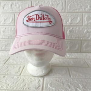 May include: A light pink trucker hat featuring a white oval patch on the front with the red embroidered text Von Dutch. The cap has a structured front panel, a curved brim with tonal stitching, and a mesh back panel in a matching pink shade.