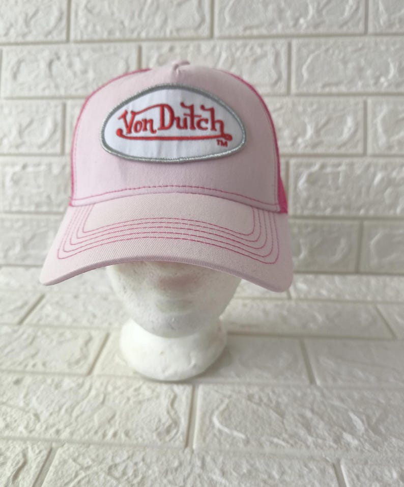 May include: A light pink trucker hat featuring a white oval patch on the front with the red embroidered text Von Dutch. The cap has a structured front panel, a curved brim with tonal stitching, and a mesh back panel in a matching pink shade.