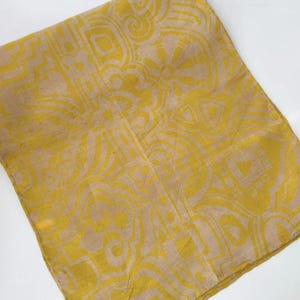 May include: A rectangular fabric scarf featuring a mustard yellow and beige geometric pattern. The design consists of repeating abstract shapes, lines, and circular motifs printed across the entire surface of the folded textile accessory.