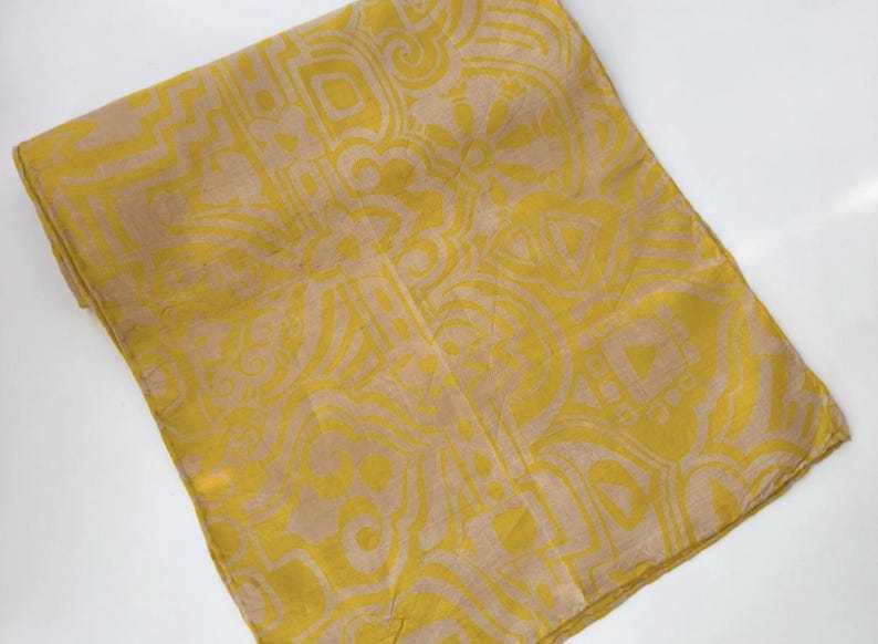 May include: A rectangular fabric scarf featuring a mustard yellow and beige geometric pattern. The design consists of repeating abstract shapes, lines, and circular motifs printed across the entire surface of the folded textile accessory.