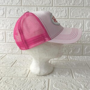 May include: A light pink trucker hat featuring a bright pink mesh back panel and a curved brim. The front panel displays a silver oval patch with red text. The hat is constructed with a structured front and an adjustable snapback closure.
