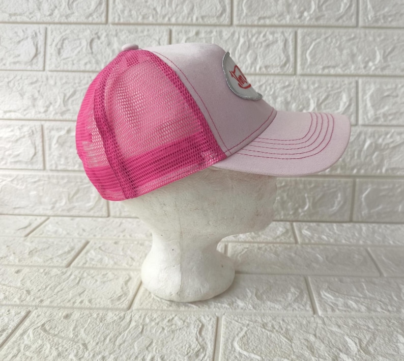 May include: A light pink trucker hat featuring a bright pink mesh back panel and a curved brim. The front panel displays a silver oval patch with red text. The hat is constructed with a structured front and an adjustable snapback closure.