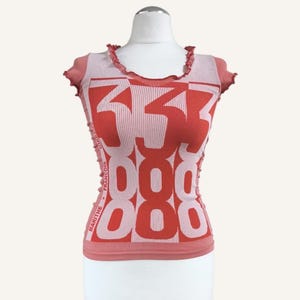 May include: A fitted, short-sleeved top in shades of pink and red. The top features a repeating pattern of large numbers and the text "MARITHE + FRANCOIS". The neckline and sleeves have a ruffled trim.