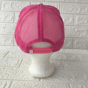 May include: A bright pink trucker hat featuring a mesh back panel and a solid front panel. The cap includes a matching pink plastic snapback closure at the rear, allowing for adjustable sizing. The design is consistent in color throughout the entire piece.