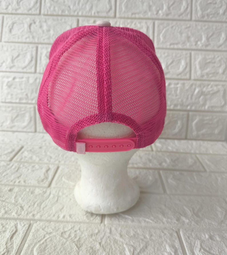 May include: A bright pink trucker hat featuring a mesh back panel and a solid front panel. The cap includes a matching pink plastic snapback closure at the rear, allowing for adjustable sizing. The design is consistent in color throughout the entire piece.