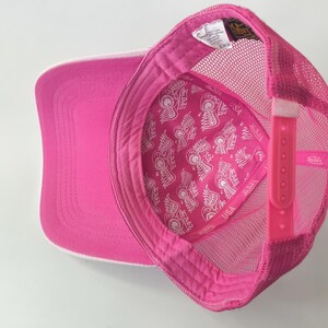 May include: A bright pink trucker hat featuring a mesh back and an adjustable plastic snapback closure. The interior lining is decorated with a repeating white graphic pattern of winged logos and the text Von Dutch USA.