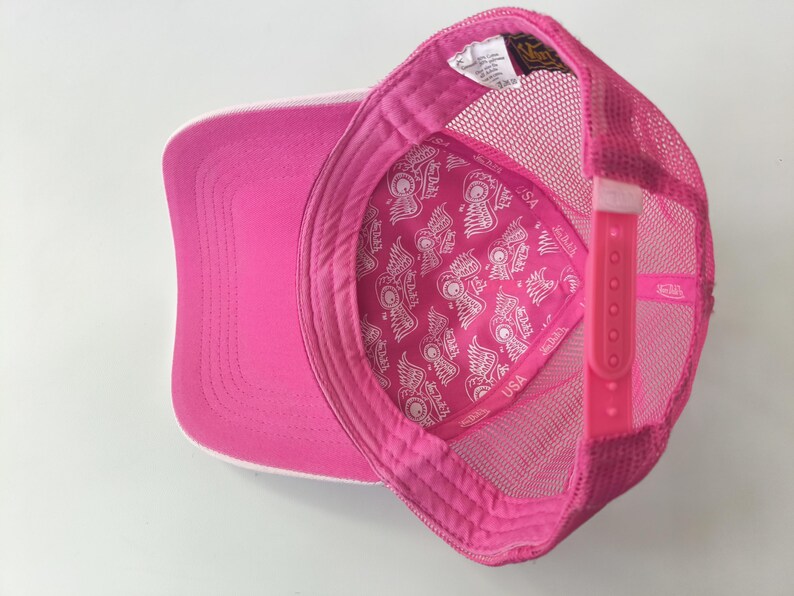 May include: A bright pink trucker hat featuring a mesh back and an adjustable plastic snapback closure. The interior lining is decorated with a repeating white graphic pattern of winged logos and the text Von Dutch USA.