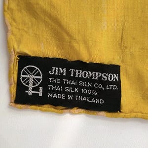 May include: A bright yellow fabric with a black rectangular label sewn onto the corner. The label features a white spinning wheel icon and text that reads: JIM THOMPSON, THE THAI SILK CO., LTD., THAI SILK 100%, MADE IN THAILAND.
