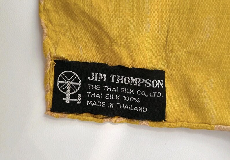 May include: A bright yellow fabric with a black rectangular label sewn onto the corner. The label features a white spinning wheel icon and text that reads: JIM THOMPSON, THE THAI SILK CO., LTD., THAI SILK 100%, MADE IN THAILAND.