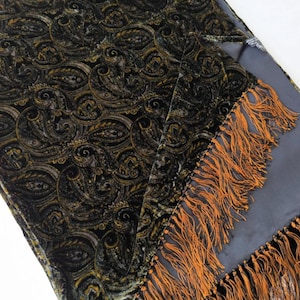 Puede incluir: Una manta de terciopelo oscuro con un estampado de cachemira en negro, dorado y verde oliva. La manta tiene un reverso gris y está adornada con largas borlas de color naranja dorado.