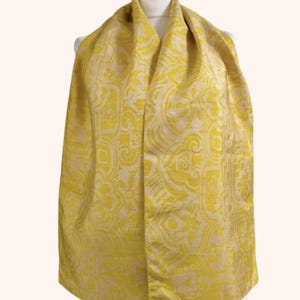May include: A bright yellow scarf featuring a repeating cream-coloured geometric and floral pattern. The scarf is draped over a mannequin, displaying a smooth, woven texture with a consistent design across the entire visible surface area.