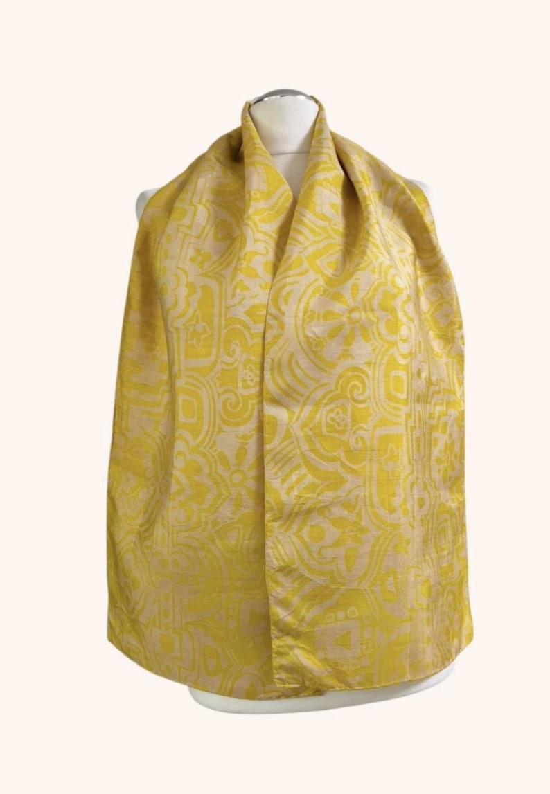 May include: A bright yellow scarf featuring a repeating cream-coloured geometric and floral pattern. The scarf is draped over a mannequin, displaying a smooth, woven texture with a consistent design across the entire visible surface area.