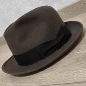 Puede incluir: Un sombrero fedora de color marrón oscuro con una copa estructurada con un pliegue central y un frente pellizcado. El sombrero está rodeado por una banda ancha de cinta de grosgrain negra que incluye un pequeño detalle de lazo en el lateral.