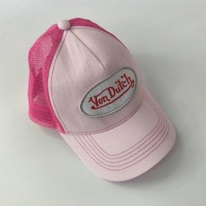May include: A light pink trucker hat featuring a mesh back in a vibrant hot pink color. The front panel is solid light pink with a central oval patch containing the red text Von Dutch. The hat includes a curved brim with matching pink stitching.