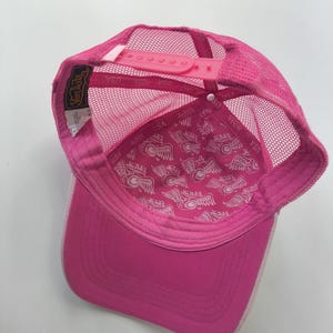 May include: A bright pink trucker hat featuring a mesh back and an adjustable plastic snapback closure. The interior lining is decorated with a repeating white Von Dutch logo pattern, and a black and gold brand label is stitched to the inner seam.