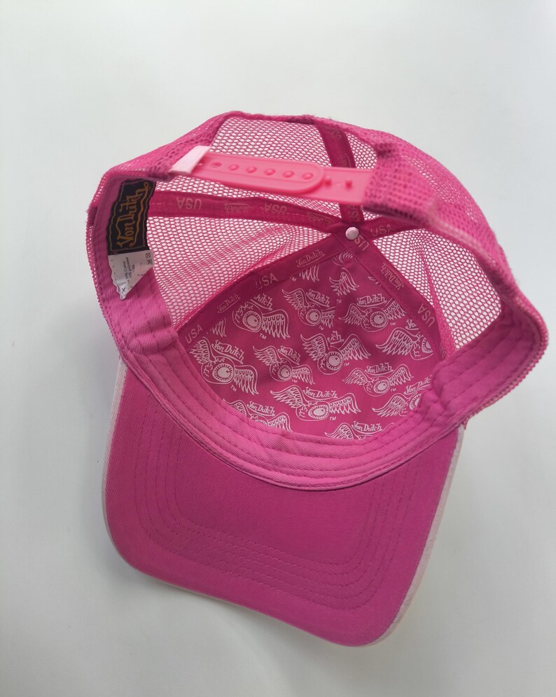 May include: A bright pink trucker hat featuring a mesh back and an adjustable plastic snapback closure. The interior lining is decorated with a repeating white Von Dutch logo pattern, and a black and gold brand label is stitched to the inner seam.