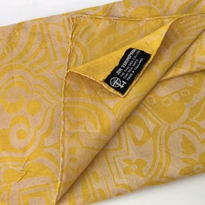 May include: A yellow and beige patterned scarf featuring a repeating abstract design. A black fabric label is sewn onto the corner with the white text JIM THOMPSON, THE THAI SILK CO., LTD, THAI SILK 100%, MADE IN THAILAND, and a small spinning wheel icon.