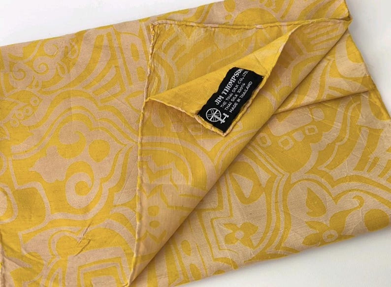 May include: A yellow and beige patterned scarf featuring a repeating abstract design. A black fabric label is sewn onto the corner with the white text JIM THOMPSON, THE THAI SILK CO., LTD, THAI SILK 100%, MADE IN THAILAND, and a small spinning wheel icon.