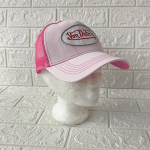 May include: A light pink trucker hat featuring a mesh back and a solid front panel. An oval patch is centered on the front with the text Von Dutch written in red script. The brim is curved and features matching pink stitching across the surface.