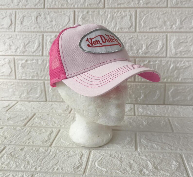 May include: A light pink trucker hat featuring a mesh back and a solid front panel. An oval patch is centered on the front with the text Von Dutch written in red script. The brim is curved and features matching pink stitching across the surface.