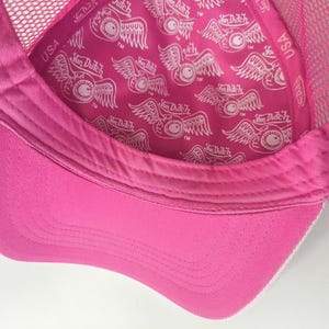 May include: A bright pink trucker hat featuring a mesh back and a solid pink brim. The interior lining is printed with a repeating white Von Dutch logo and a winged eyeball graphic. The inner sweatband also displays the Von Dutch brand name and USA.