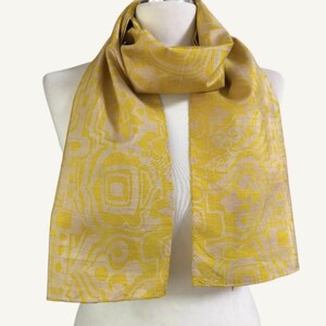 May include: A rectangular scarf featuring a vibrant mustard yellow and light beige abstract geometric pattern. The fabric displays a subtle sheen with a woven texture, draped around a mannequin neck, showcasing a repeating design of shapes.