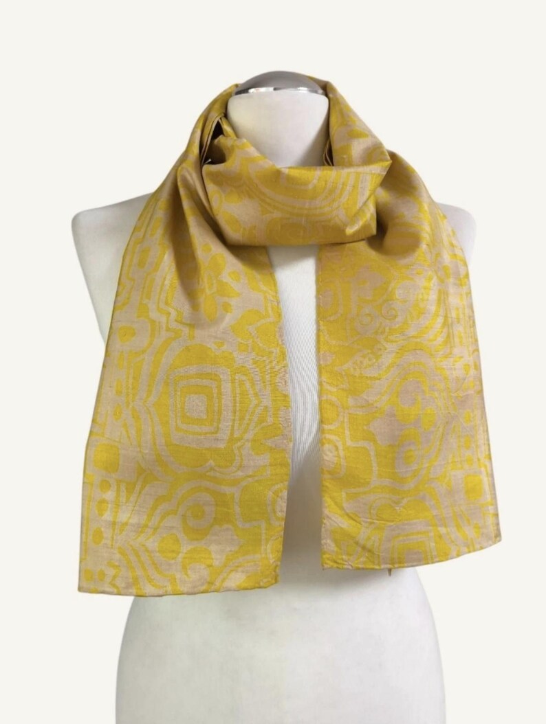 May include: A rectangular scarf featuring a vibrant mustard yellow and light beige abstract geometric pattern. The fabric displays a subtle sheen with a woven texture, draped around a mannequin neck, showcasing a repeating design of shapes.