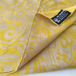 May include: A yellow and beige fabric featuring a repeating geometric pattern. A black rectangular tag is sewn into the corner, displaying the text JIM THOMPSON, THE THAI SILK CO. LTD, THAI SILK 100%, MADE IN THAILAND, and a small circular logo.