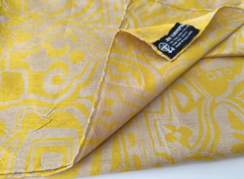 May include: A yellow and beige fabric featuring a repeating geometric pattern. A black rectangular tag is sewn into the corner, displaying the text JIM THOMPSON, THE THAI SILK CO. LTD, THAI SILK 100%, MADE IN THAILAND, and a small circular logo.